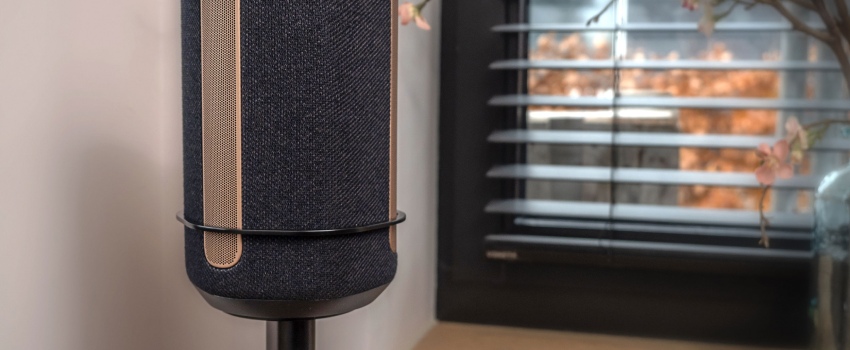 Primer plano de un altavoz negro sobre un soporte Vebos con un soporte, colocado en un interior moderno junto a una ventana con persianas.